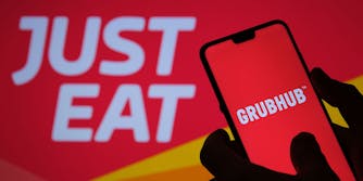 GrubHub app screen on black phone held in hand silhouette with "Just Eat" blurred in red and yellow background
