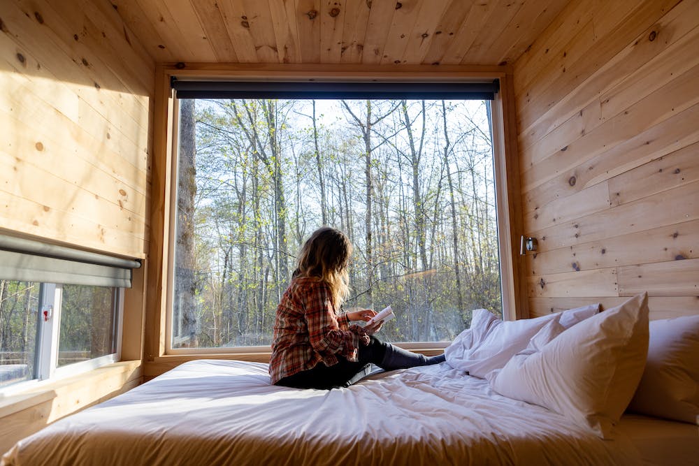 a woman with tattoos sitting on a bed inside a getway houes cabin reading a book