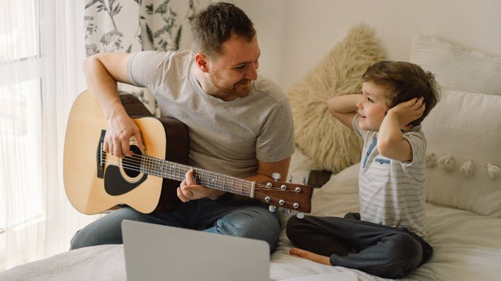 Father and son learn to play the acoustic guitar in an online lesson. Free time to spend with my son at home, teaching him to play the guitar.