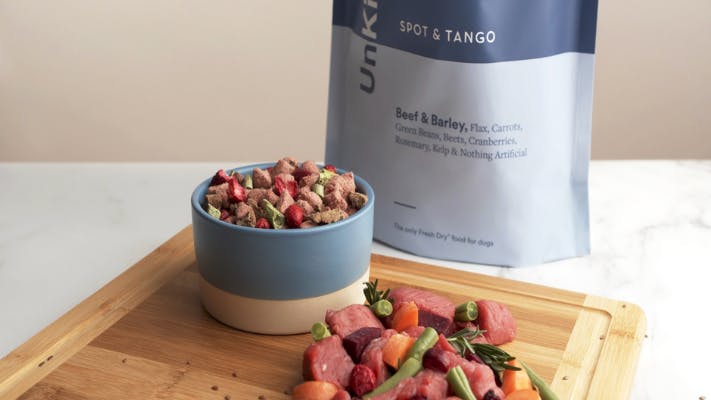 Bowl of Spot and Tango fresh dog food on a wooden cutting board next to raw meats and vegetables