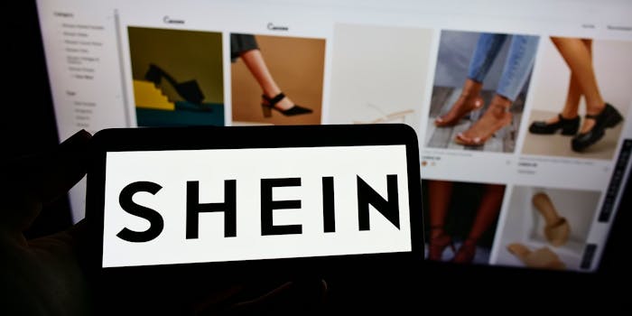 silhouette of hand holding phone with SHEIN logo on screen in front of an open laptop on SHEIN website