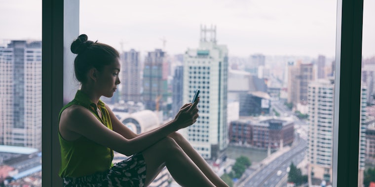 Woman staying home using mobile phone by window indoors in the dark. Asian young lady pensive looking at cellphone social media app. Mental health online addiction during covid confinement concept.