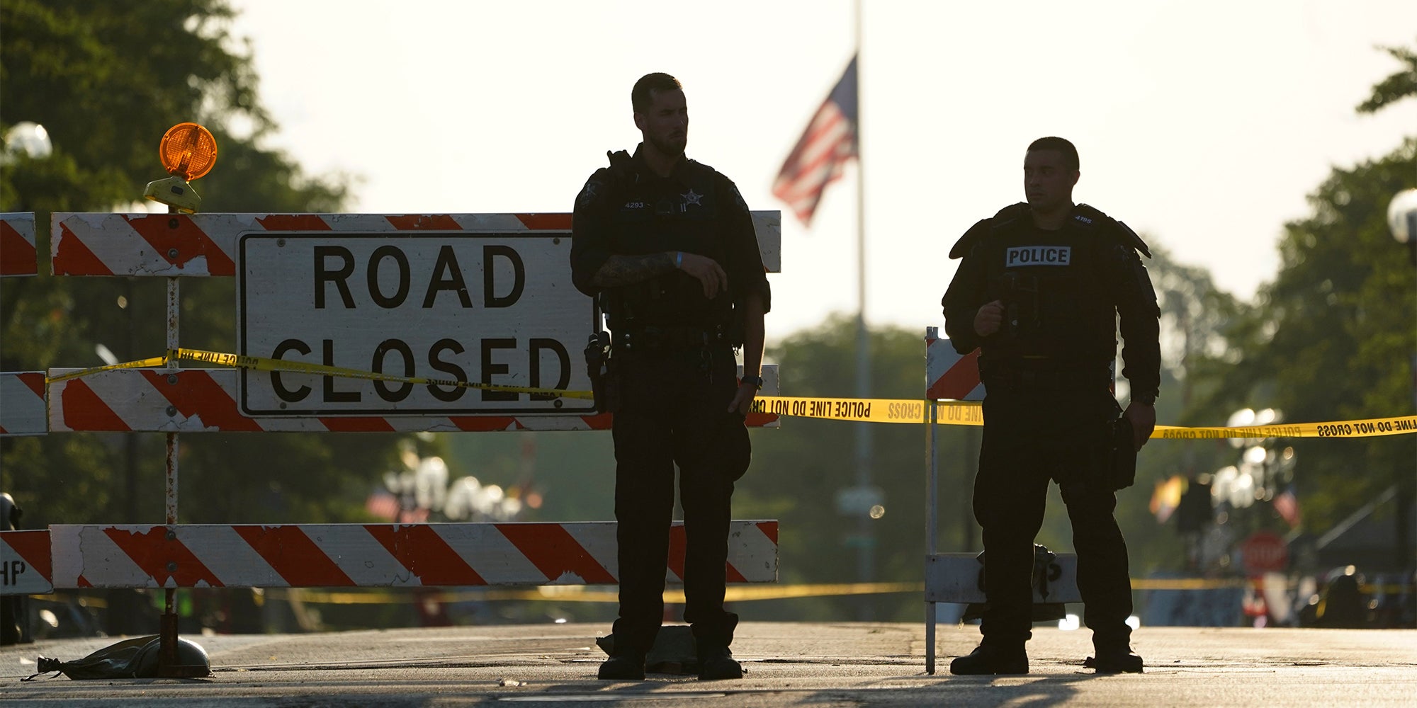 Two police officers stand their post, the day after a deadly mass shooting, on the Westside of the Highland Park, Ill., as the American flag flies at half-staff on the Eastside. A shooter fired on an Independence Day parade from a rooftop spraying the crowd with gunshots initially mistaken for fireworks before hundreds of panicked revelers of all ages fled in terror