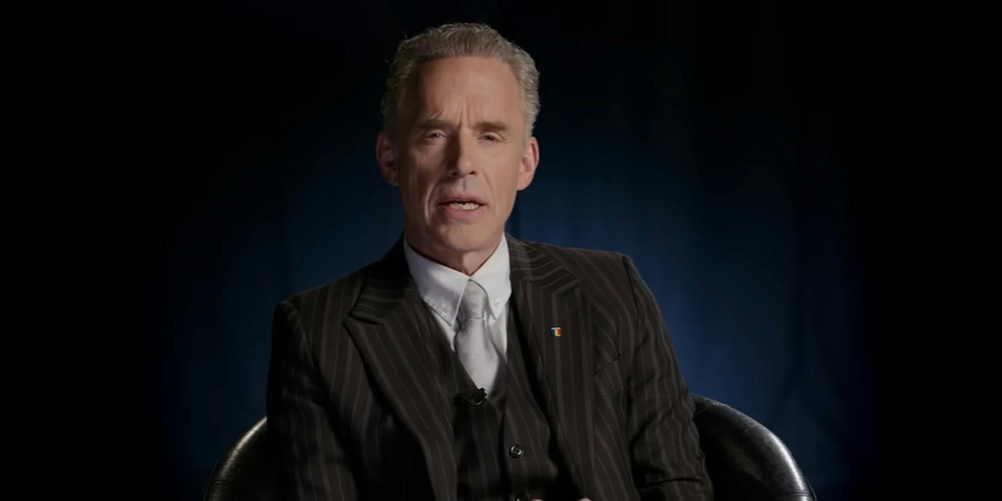 Jordan Peterson sitting in chair speaking on dark blue background (c)