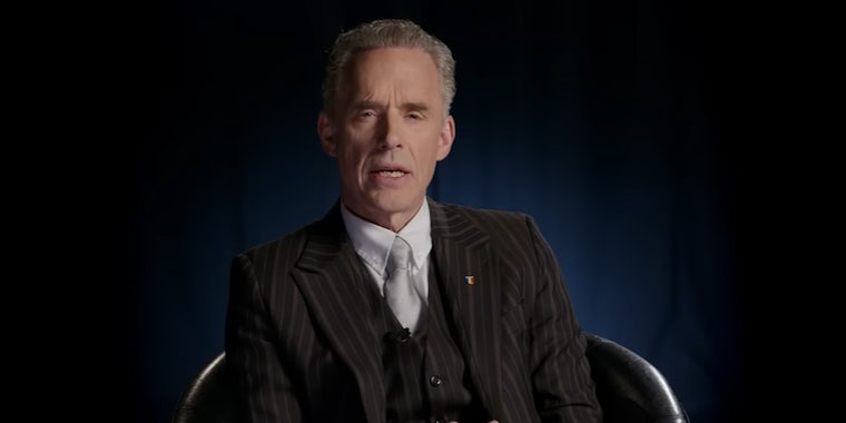 Jordan Peterson sitting in chair speaking on dark blue background (c)