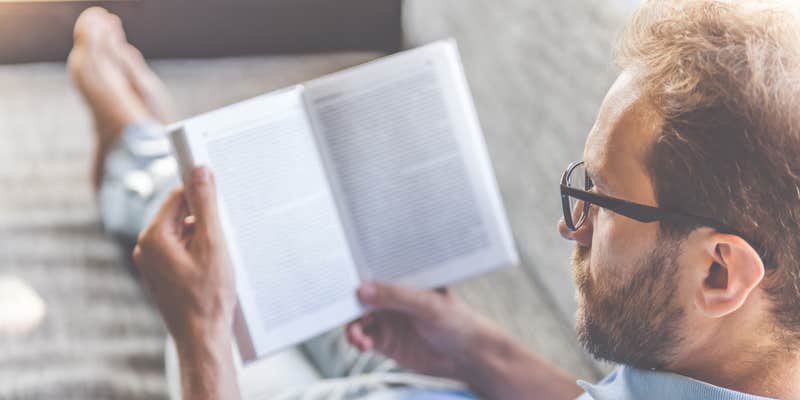 Back view of young man in casual clothes and eyeglasses reading a book while lying on couch at home