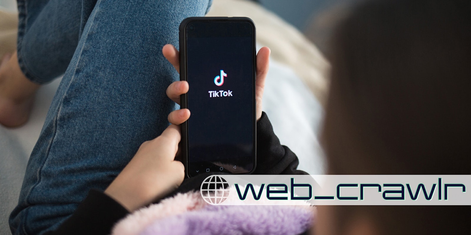 A woman holding a phone with the TikTok logo on it. In the bottom right corner is the web_crawlr Daily Dot newsletter logo.