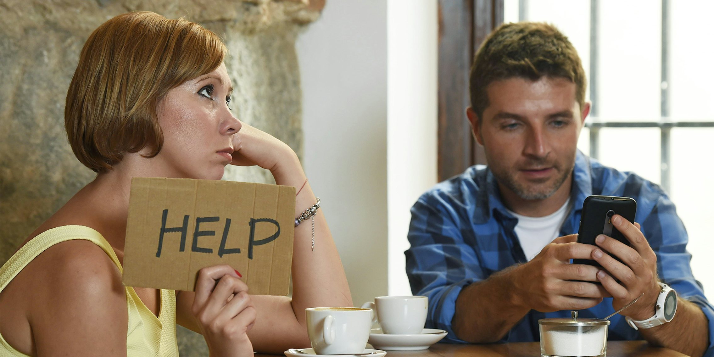 woman holding 'help' sign while man looks at phone