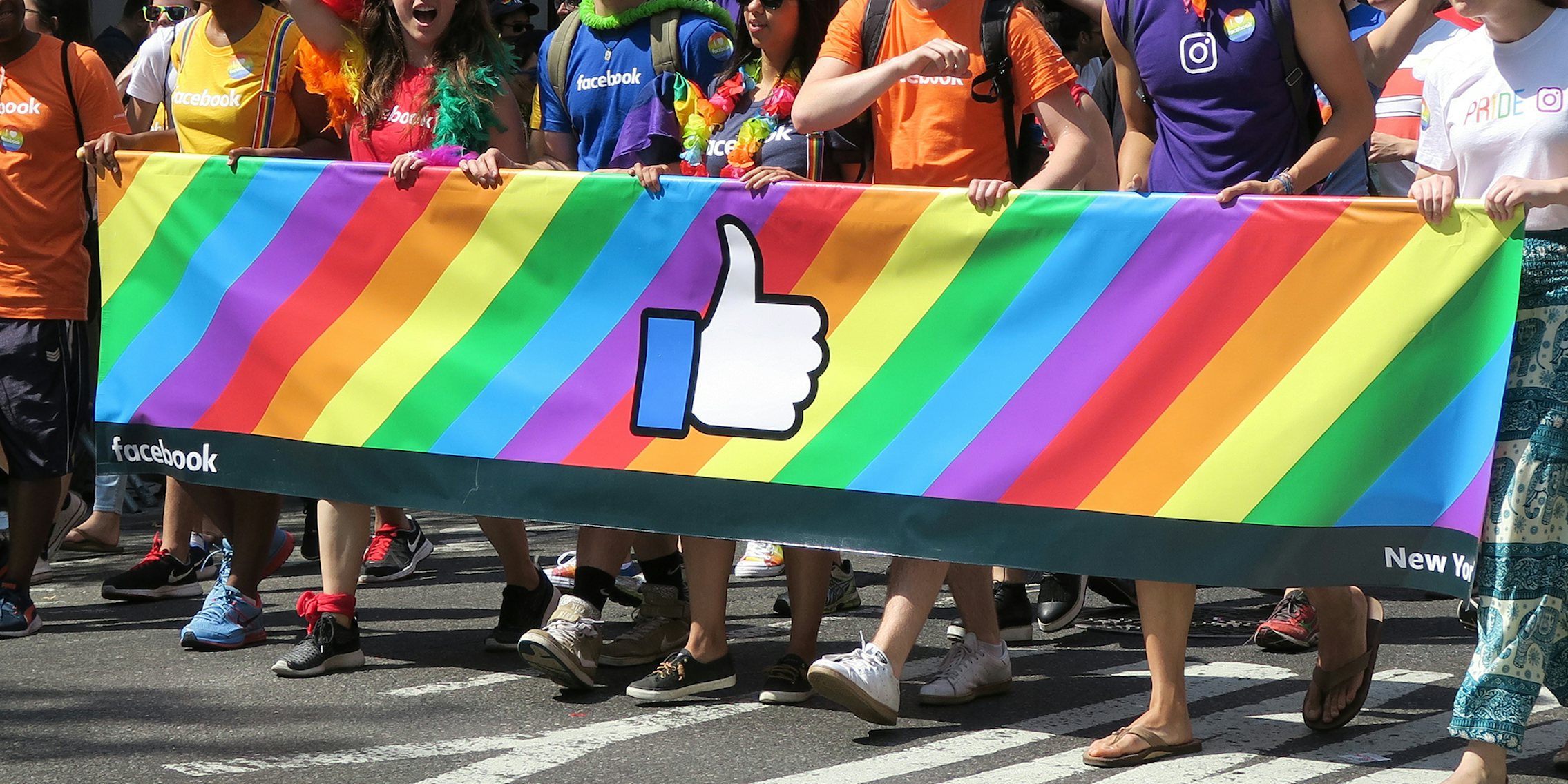 Group of people in the Gay Pride Parade behind a rainbow colored banner with the Facebook logo on it.