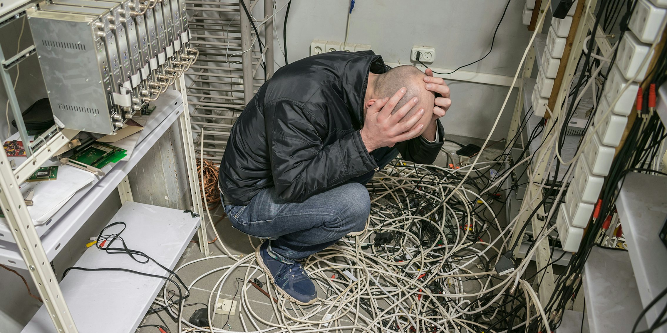 A sad technician sits near a pile of wires and holds his head with his hands in an empty datacenter. The man is in a plundered server room.