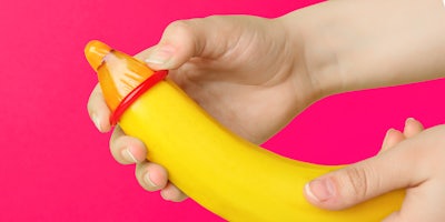 Woman putting condom on banana against color background