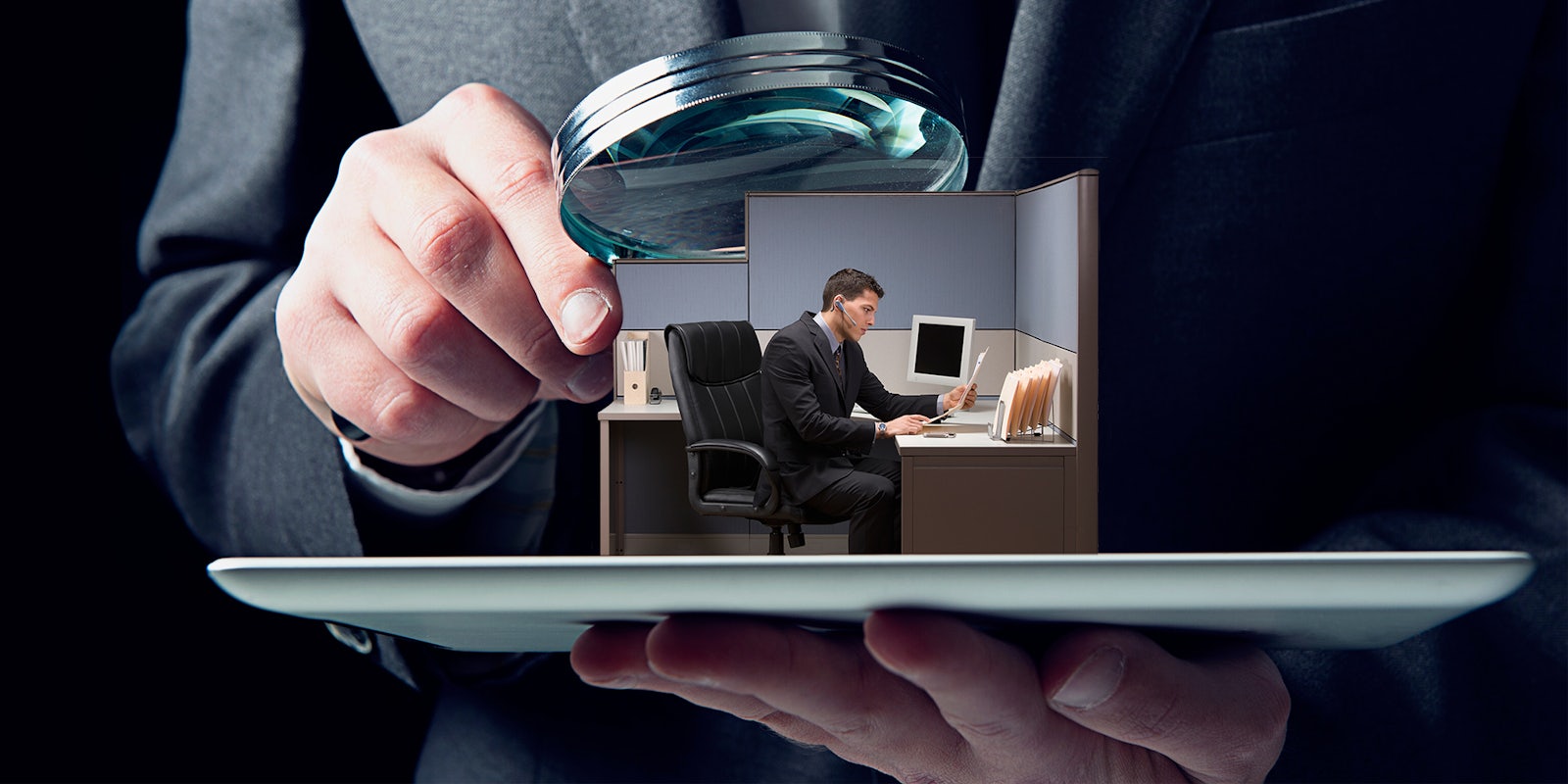 small businessman in cubicle on tablet being examined by large man holding magnifying glass