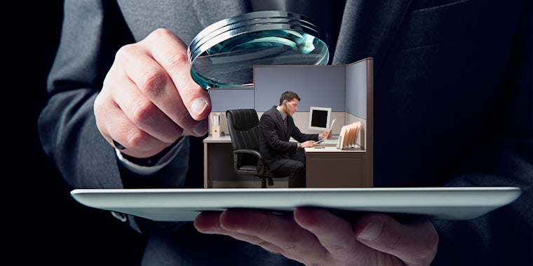 small businessman in cubicle on tablet being examined by large man holding magnifying glass