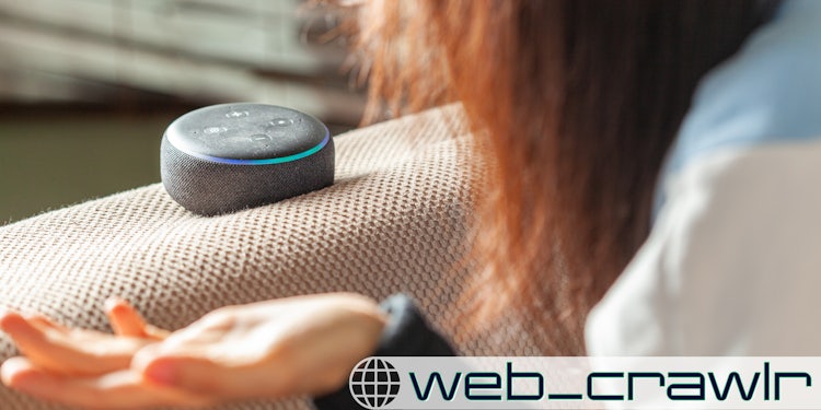 A woman gesturing at an Amazon Alexa device. The Daily Dot newsletter web_crawlr logo is in the bottom right corner.