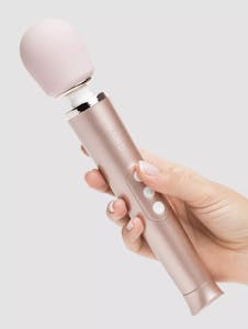 A hand holding a rose gold Le Wand Petite vibrator against a grey background.
