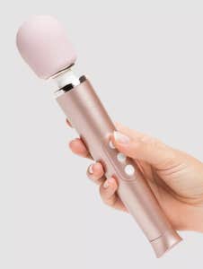 A hand holding a rose gold Le Wand Petite vibrator against a grey background.