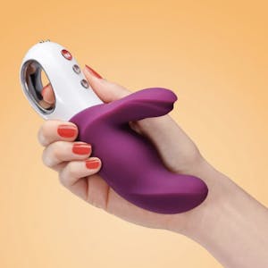 A woman's hand holding a plum-colored rabbit-style Miss Bi vibrator against an orange background.