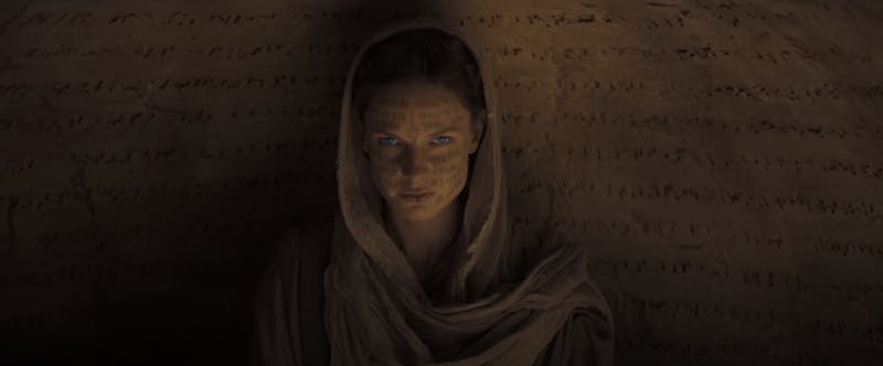 A screenshot from the Dune trailer showing Lady Jessica against a backdrop of ancient-looking text, which is also projected over her face.