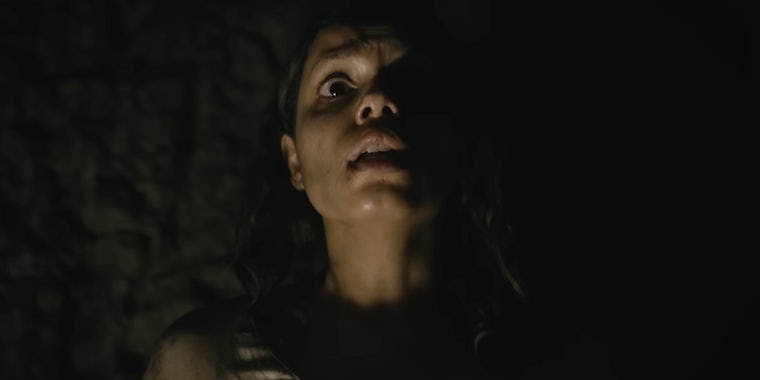 A screenshot from the Barbarian (2022) trailer showing a frightened young woman with her back against a wall and her face partially illuminated.
