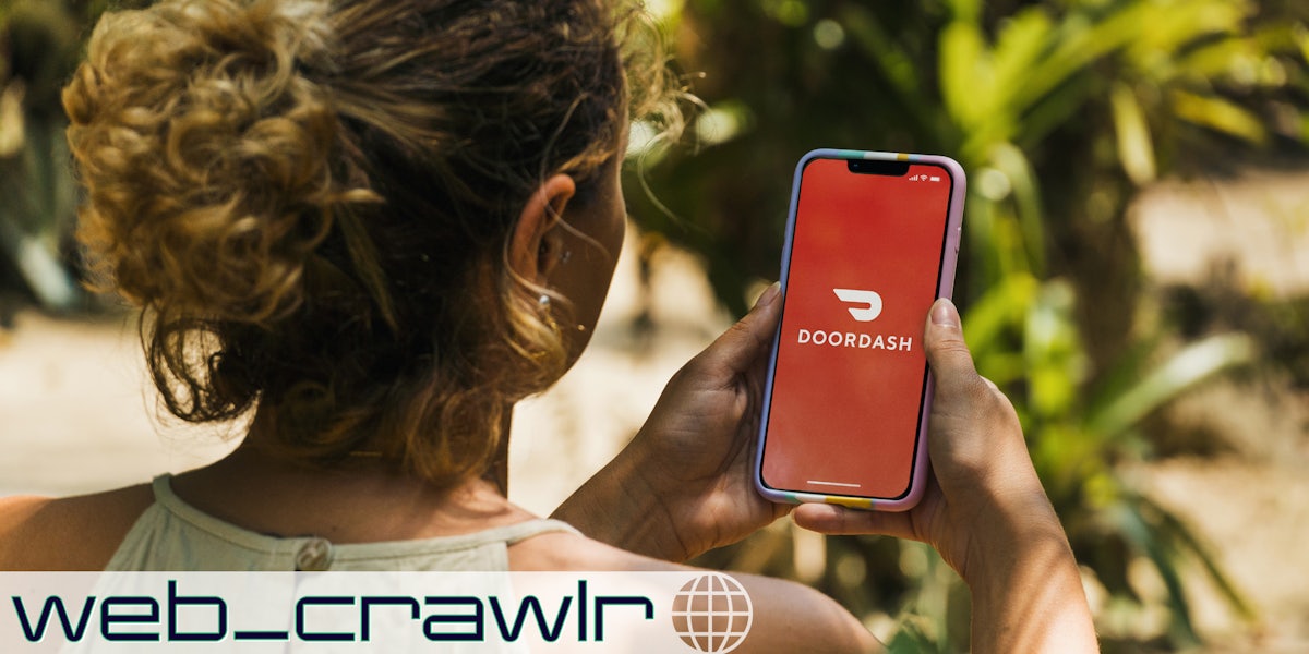 A woman holding a smartphone with the DoorDash logo on it. The Daily Dot newsletter web_crawlr logo is in the bottom left corner.