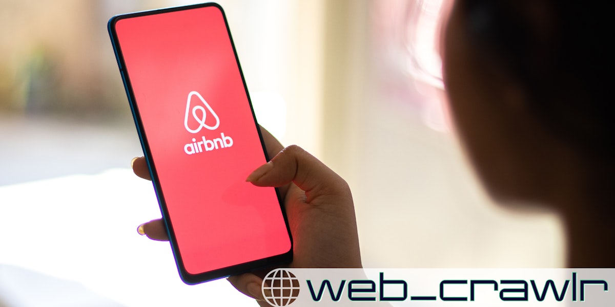 A woman holding a phone with the Airbnb logo on it. The Daily Dot newsletter web_crawlr logo is in the bottom right corner.