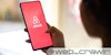 A woman holding a phone with the Airbnb logo on it. The Daily Dot newsletter web_crawlr logo is in the bottom right corner.