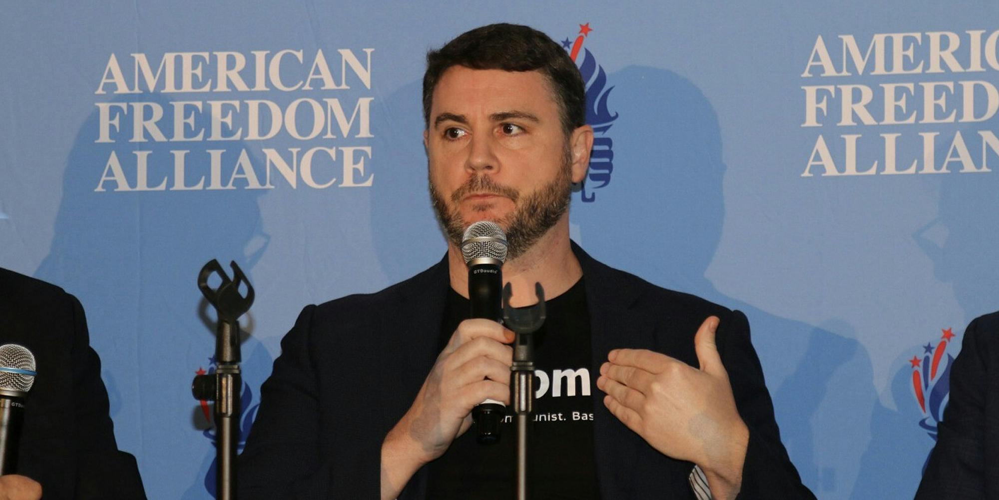 James Lindsay speaking in microphone in front of blue "AMERICAN FREEDOM ALLIANCE" background