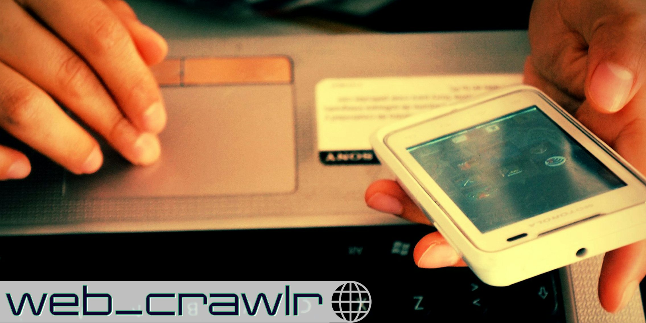 A person holding a smartphone on a laptop. The Daily Dot newsletter web_crawlr logo is in the bottom left corner.