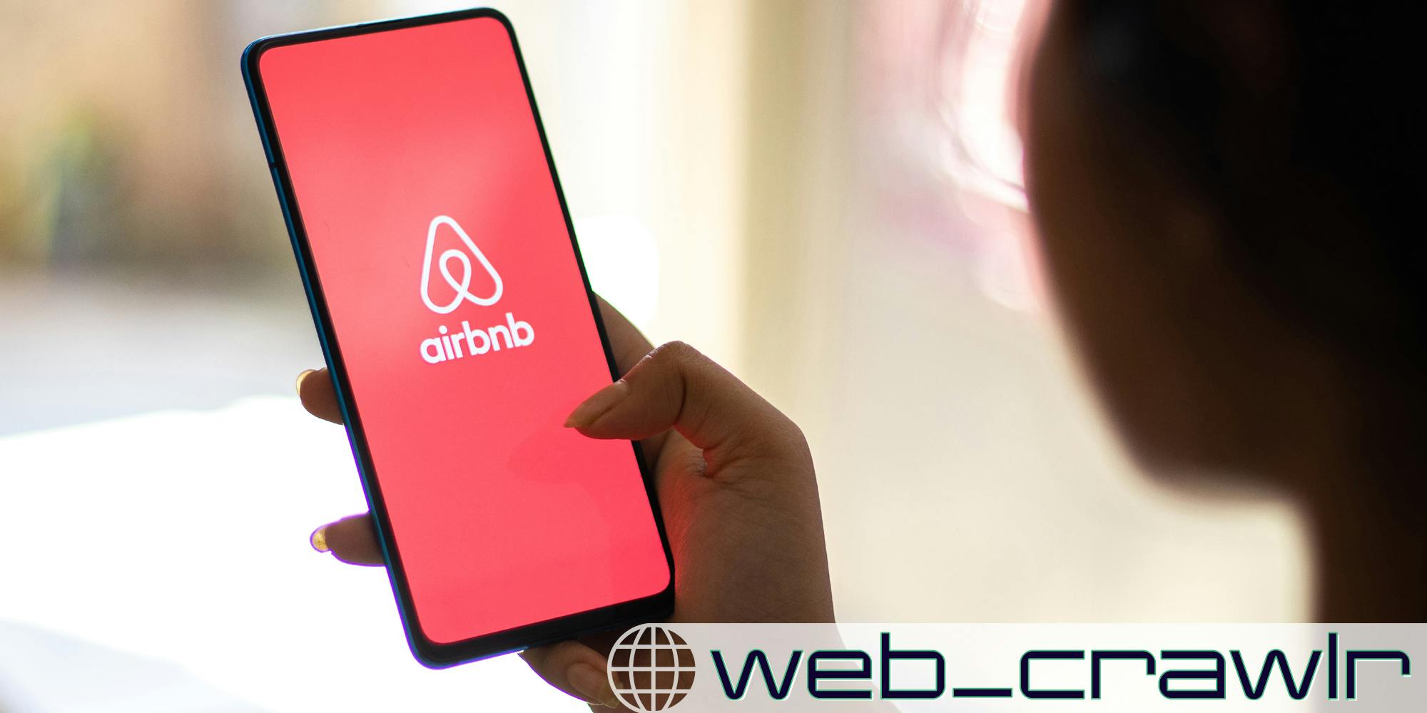 A person holding a smartphone with the Airbnb logo on it. The Daily Dot newsletter web_crawlr logo is in the bottom right corner.