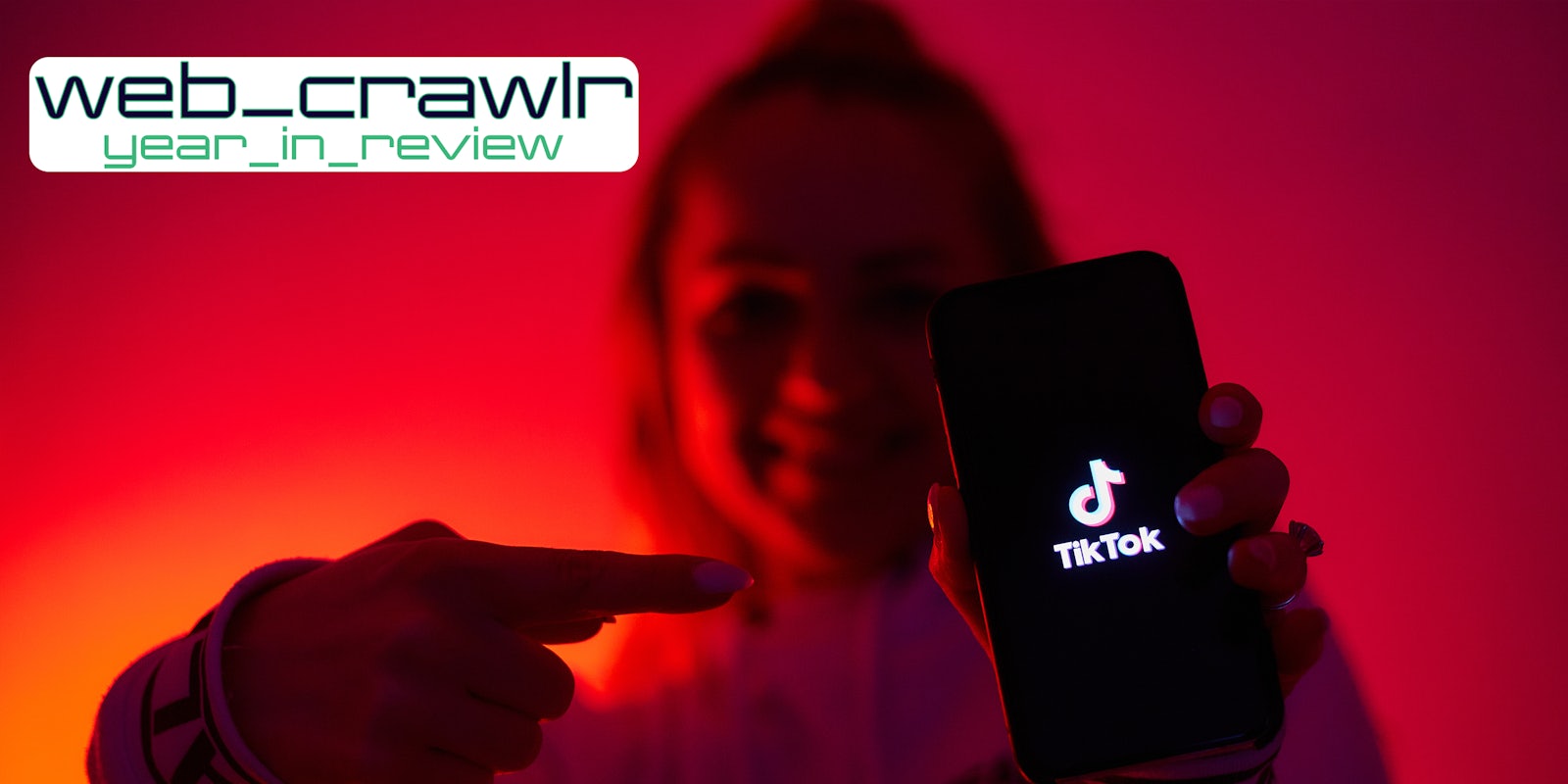 A person holding a phone with the TikTok logo on it while pointing at it. The Daily Dot newsletter web_crawlr logo is in the top left corner.
