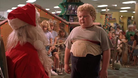 A chubby young boy with his underwear pulled up out of his pants standing in front of Santa.