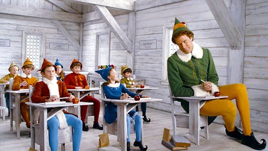 best Christmas movies streaming - Will Ferrell dressed as an elf sitting in a white classroom at a comically. small desk beside much smaller elves at desks.