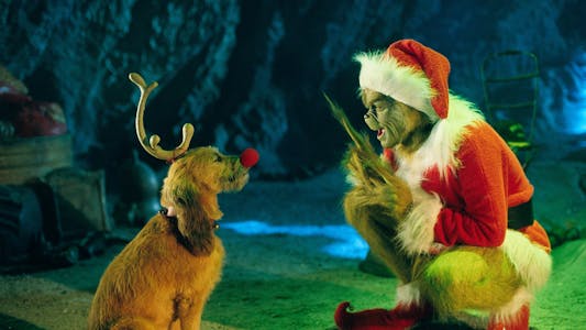 Jim Carrey as The Grinch in a Santa Claus costume crouching down and talking to his dog who has a fake red nose and an antler tied to his head.
