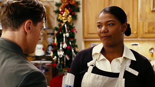 Queen Latifah wearing an apron in a kitchen decorated for Christmas looking at a man.
