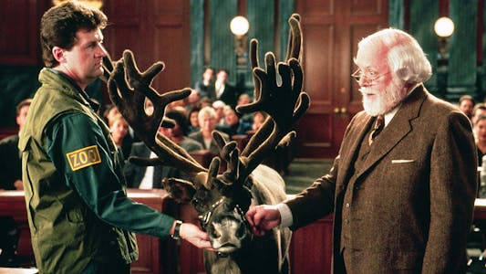 best Christmas movies streaming - A zookeeper and a man who looks like Santa Claus in a brown suit both touching a reindeer between them in a courtroom.