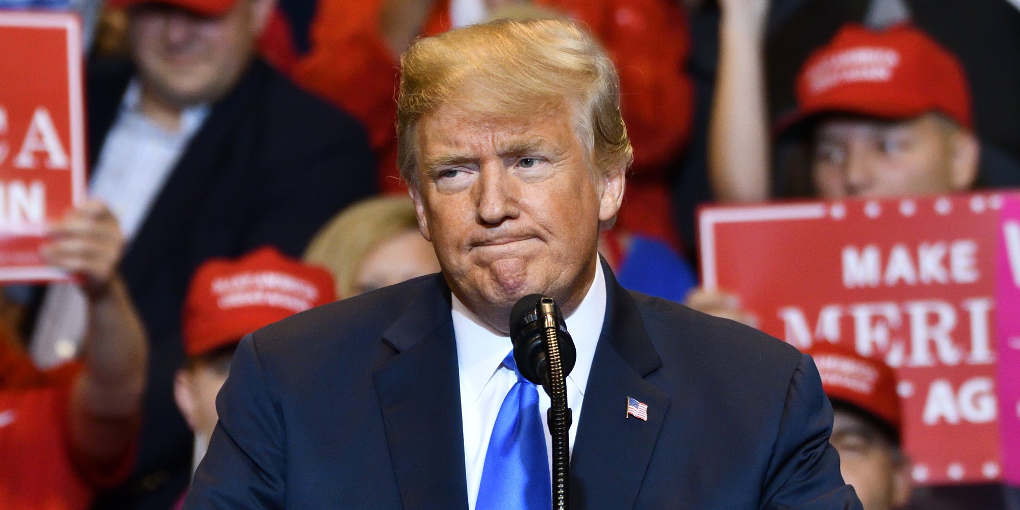 Donald Trump frowning at microphone with supporters holding signs behind