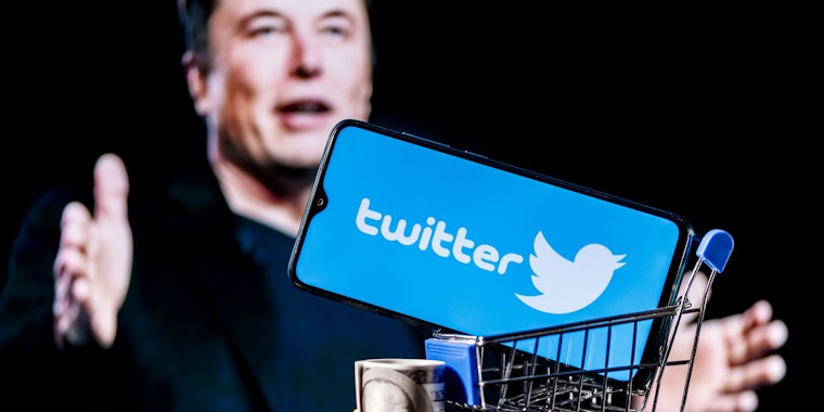 Elon Musk speaking with hands out with shopping cart containing phone with Twitter logos on screen in front of black background