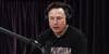 Elon Musk speaking into microphone with headphones on in front of black fabric background