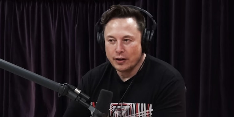 Elon Musk speaking into microphone with headphones on in front of black fabric background