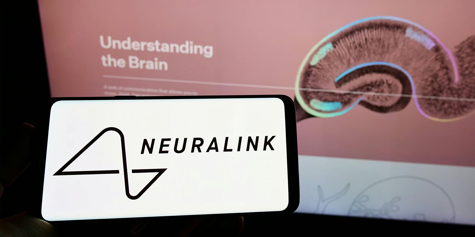Neuralink website on monitor with hand holding phone with "Neuralink" on screen in front