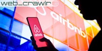 A person holding a phone with the Airbnb logo on it. The Daily Dot newsletter web_crawlr logo is in the top left corner.