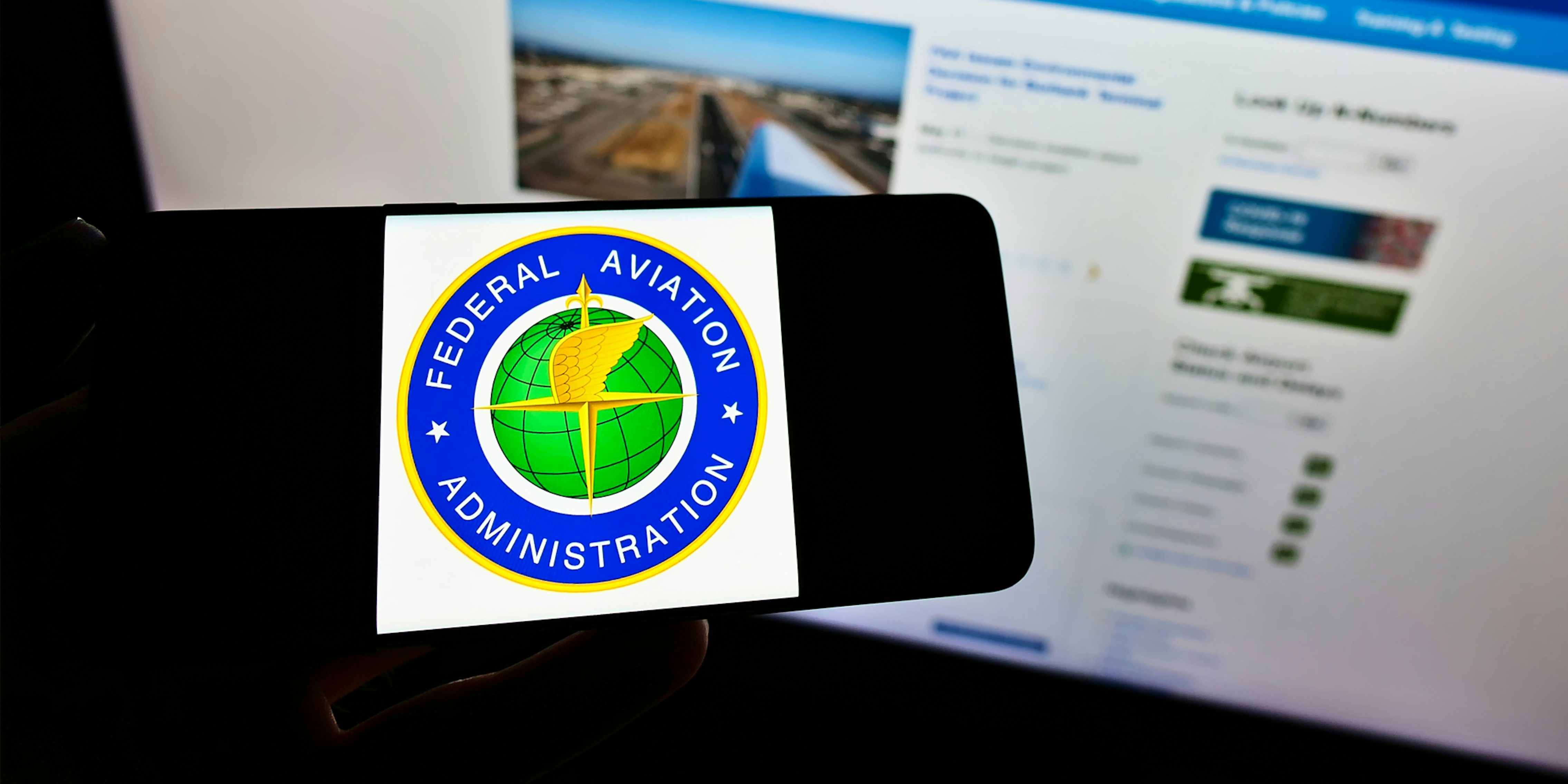 Federal Aviation Administration on phone in hand in front of monitor with FAA website on screen