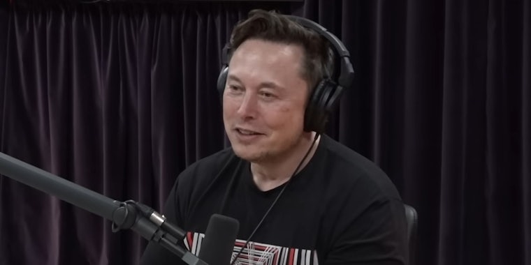 Elon Musk smiling with headphones and microphone in front of black fabric background