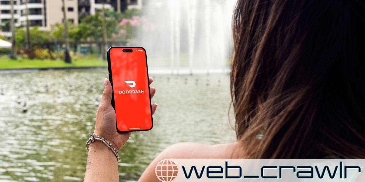 A woman holding a smartphone with the DoorDash logo on it. The Daily Dot newsletter web_crawlr logo is in the bottom right corner.