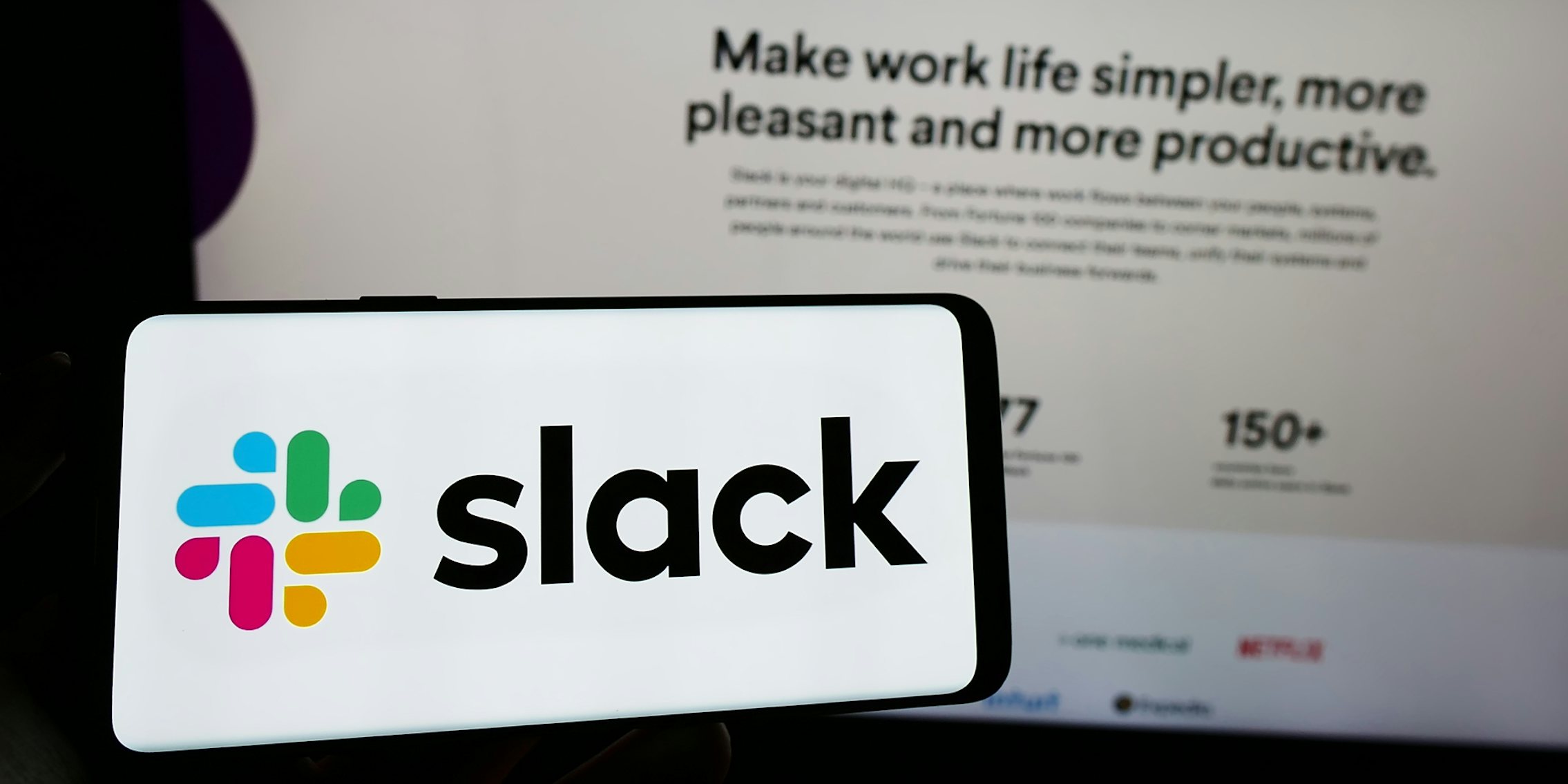 hand holding phone with Slack on screen in front of computer screen with Slack website