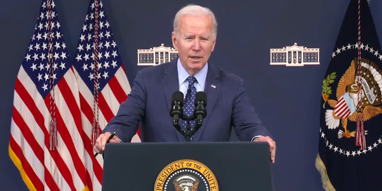 President Joe Biden speaking into microphones in front of blue background with flags at both of his sides