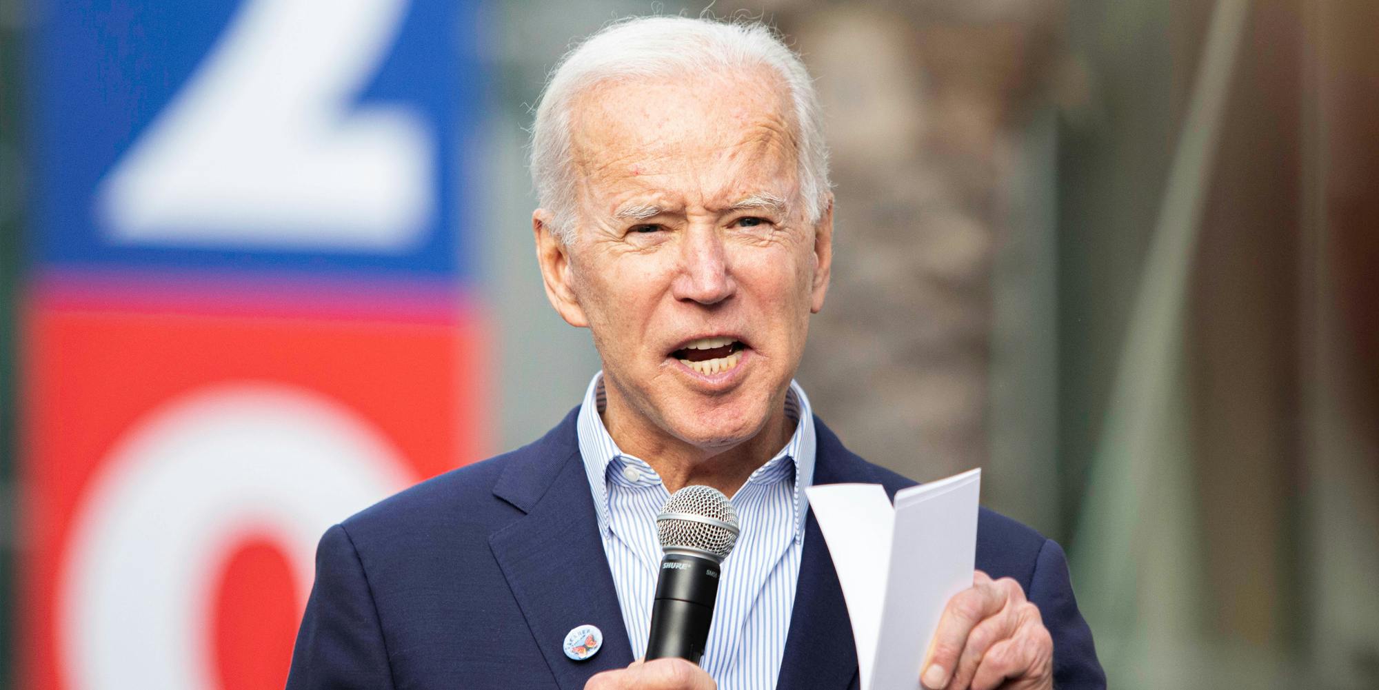 Joe Biden speaking into microphone and holding paper in front of blurred background