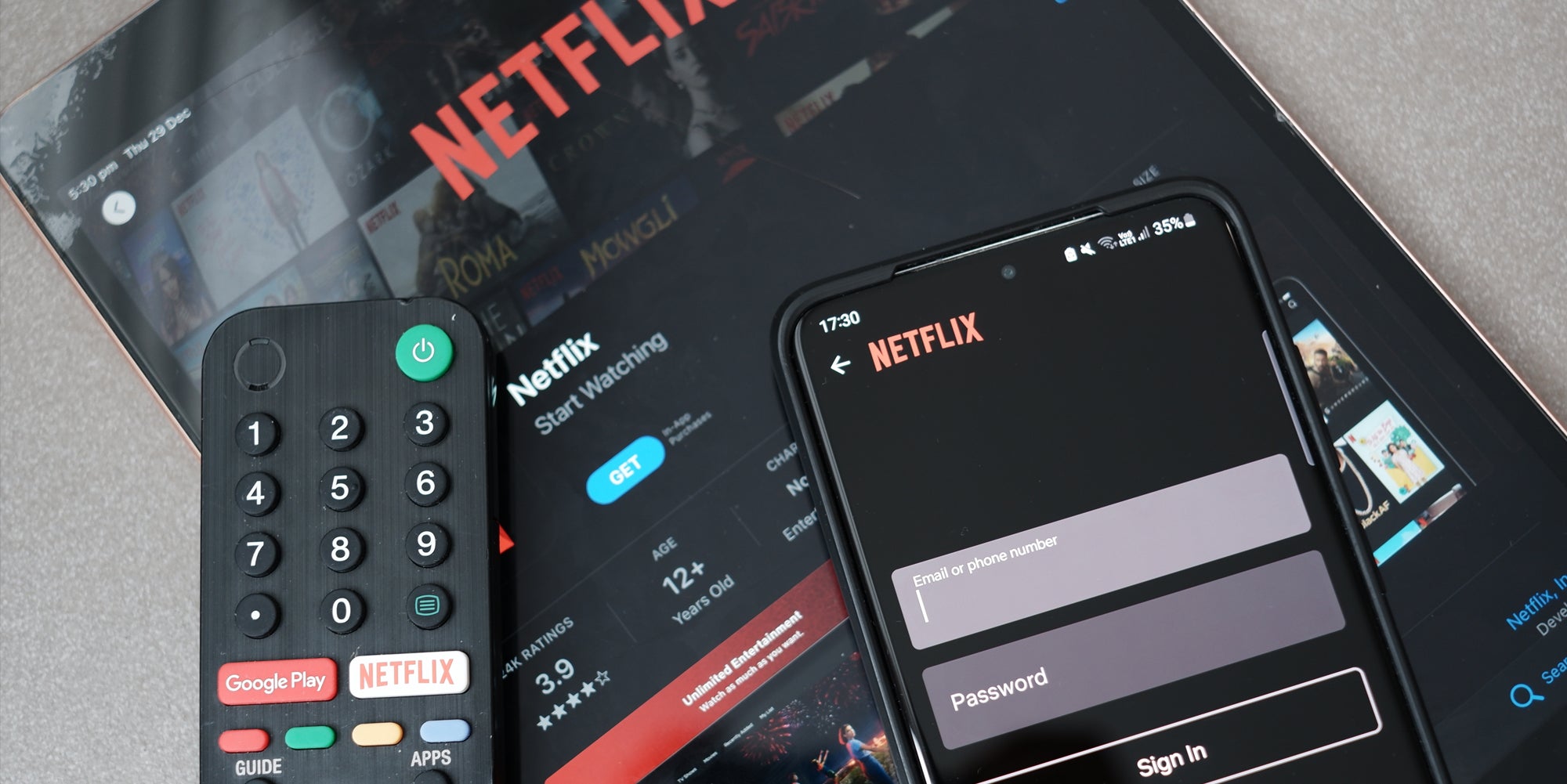 Netflix on tablet with phone on Netflix login on top next to tv remote on light grey background