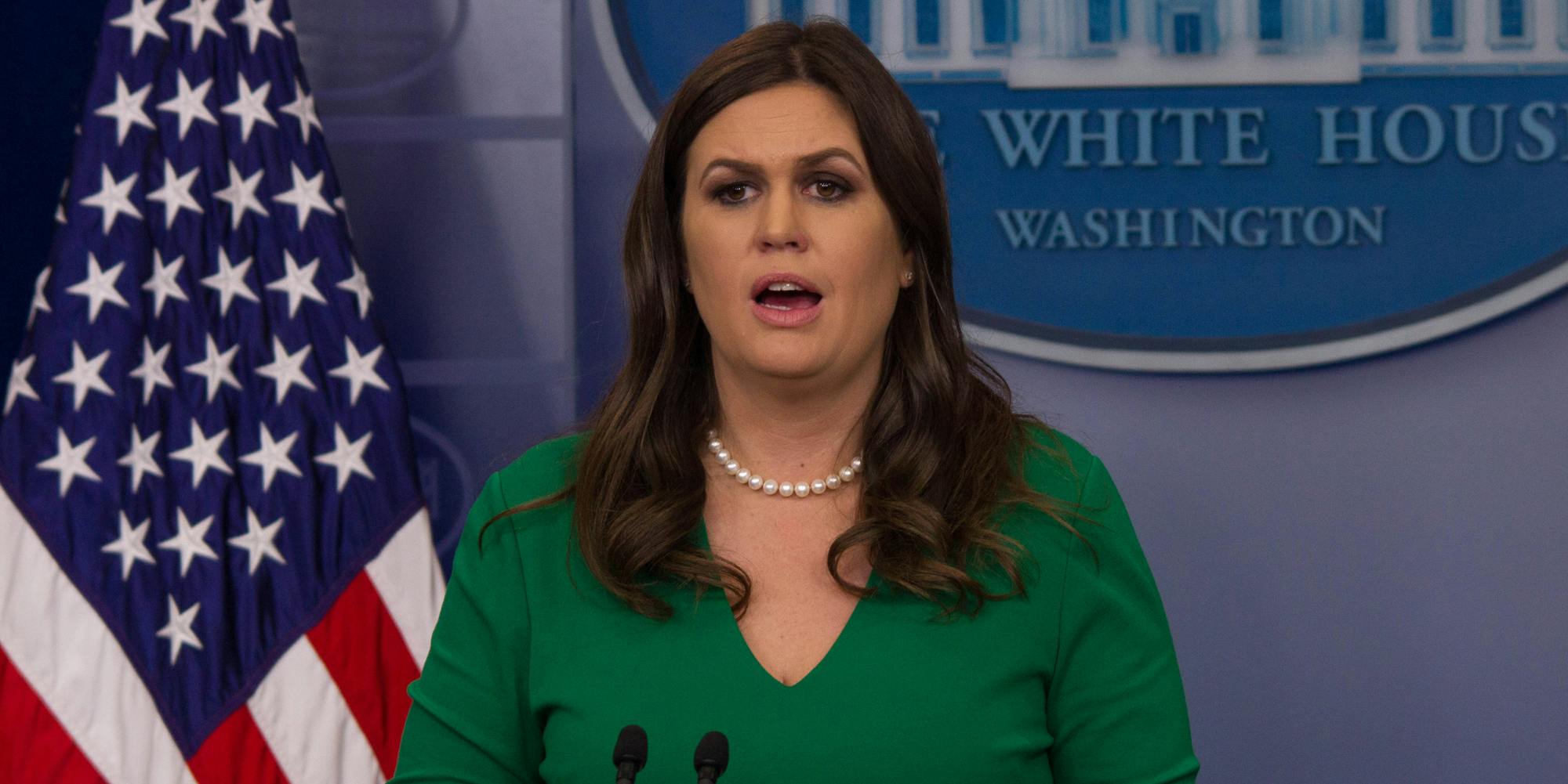 Sarah Huckabee Sanders speaking into microphones in front of blue background and American flag