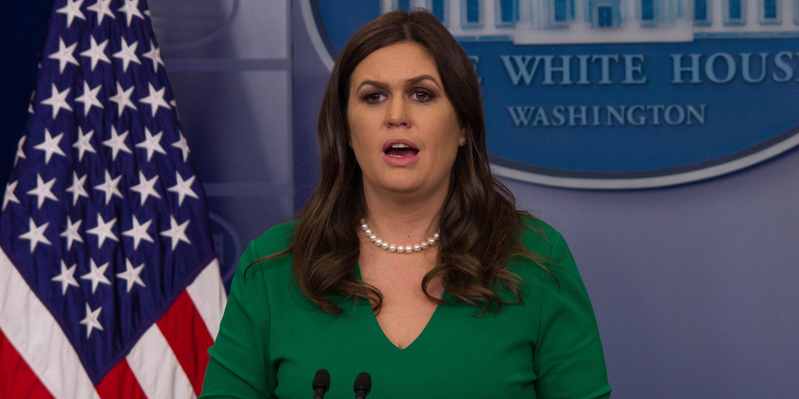 Sarah Huckabee Sanders speaking into microphones in front of blue background and American flag